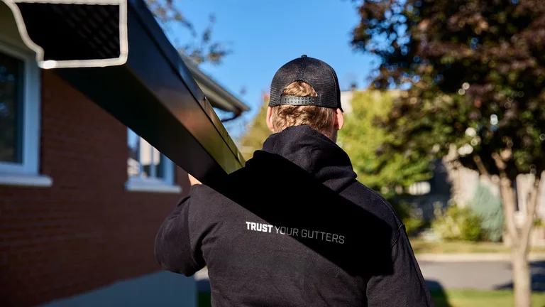Installer carrying a dark seamless gutter section toward a Rochester-area home